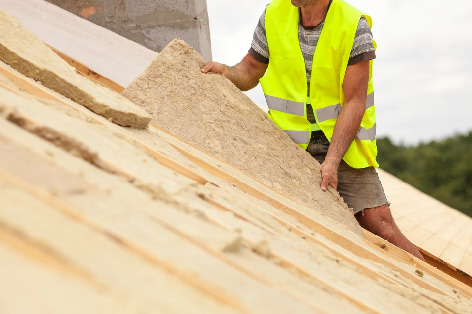 Chantier de construction extérieur avec ouvrier en bleu de travail et casque jaune installant un panneau isolant sur façade depuis échafaudage mobile